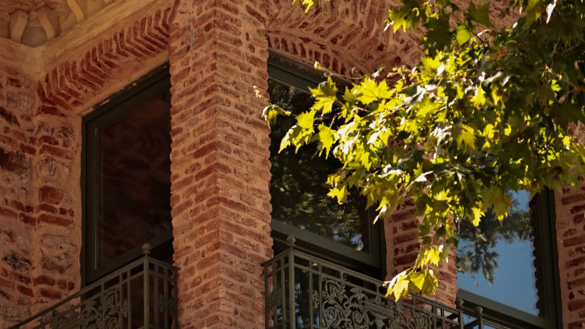 A sunlit brick building corner with intricate railing and vibrant green leaves.